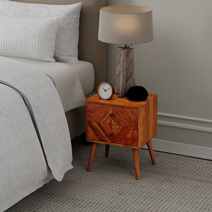 Wooden bedside table with a lamp, small clock, and a round black object. Its next to a bed with white bedding on a carpeted floor against a light-colored wall.
