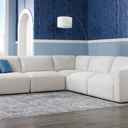 Modern living room with a large, light gray sectional sofa, a blue patterned accent wall, and a standing floor lamp. A dark blue and white rug lies on a dark wood floor. A window lets in natural light.