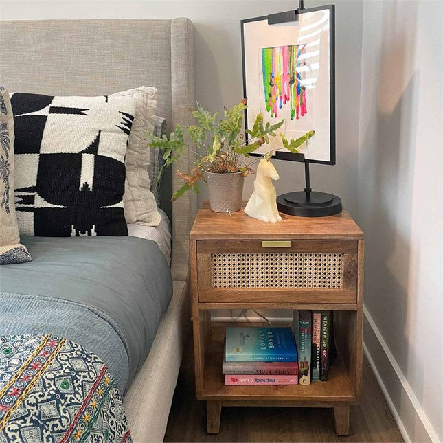 A serene bedroom scene features the rustic Bedside Table 1 Drawer with a woven cane panel beside the gray upholstered bed. It holds a plant, abstract art, and a decorative object, while books are neatly stacked on its lower shelf. The bedding displays dark and light geometric patterns.