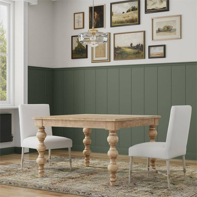 A dining room showcases a French Country Square 48 Dining Table made from solid mango wood with two white upholstered chairs on an ornate rug. The green half-painted walls display framed artworks, while a modern chandelier hangs above and a large window is to the left.