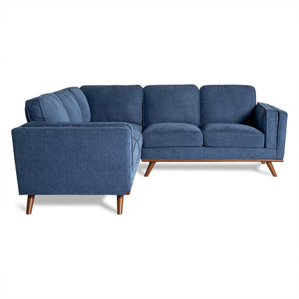 The Maddox Sectional is a blue L-shaped sofa perfect for living rooms, featuring clean lines and mid-century modern wooden tapered legs. It offers spacious seating with each side having three cushions set against a plain white backdrop.