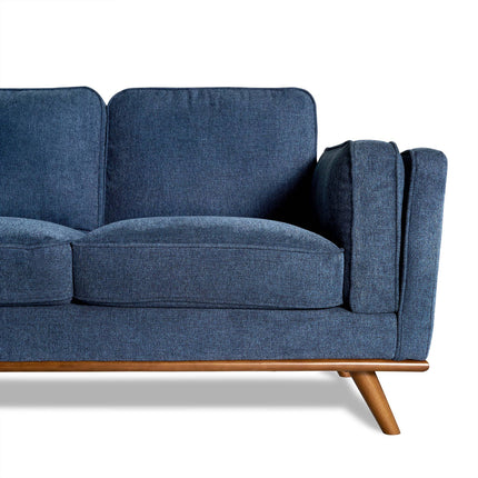 The Maddox Sectional, with its blue fabric upholstery and mid-century modern style, features wooden legs and armrests. Set against a white background, its plush cushions bring elegance to any living room. The focus is on the right half of this minimalist design.