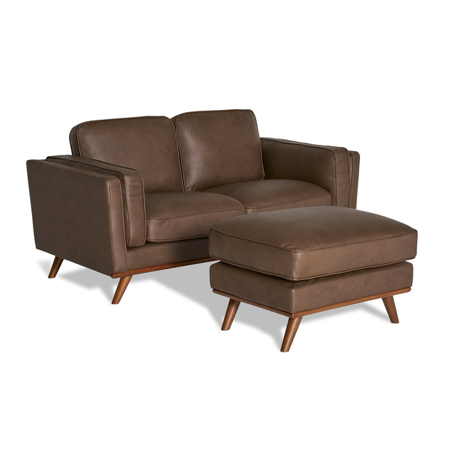 The Maddox Loveseat is a brown leather, mid-century modern piece with angled armrests and wooden legs. It includes a matching ottoman and features durable construction with two cushions on the seat and backrest for enhanced comfort.
