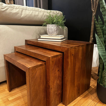 Beside a beige sofa, a set of three Acacia Nesting Tables is arranged. The top table holds stacked books and a potted plant. Ideal for small spaces, the room features a wooden floor with a tall plant to the right.