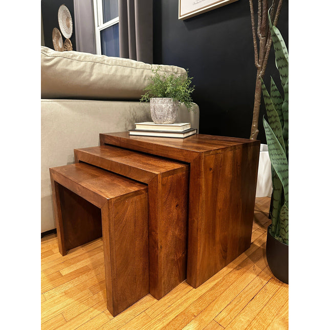 Beside a beige sofa, a set of three Acacia Nesting Tables is arranged. The top table holds stacked books and a potted plant. Ideal for small spaces, the room features a wooden floor with a tall plant to the right.