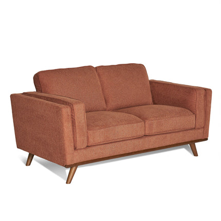 The Maddox Loveseat offers a mid-century modern style in warm rust color, featuring wooden legs and durable construction. It includes two large seat and back cushions, wide armrests, and a slightly reclined backrest, all set against a plain background.